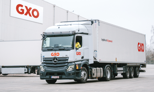 GXO-branded delivery truck at a logistics facility, with GXO signage visible on the warehouse in the background.